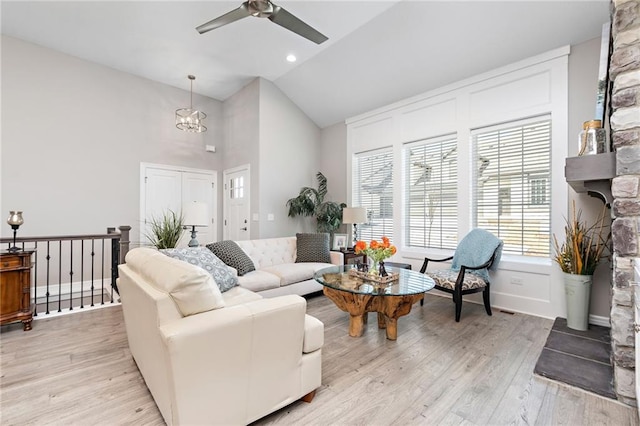 living room with high vaulted ceiling, ceiling fan with notable chandelier, a stone fireplace, and light hardwood / wood-style flooring