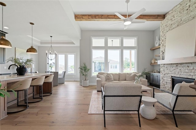 living room with ceiling fan with notable chandelier, light hardwood / wood-style floors, beamed ceiling, and a stone fireplace