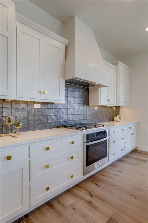 kitchen featuring custom exhaust hood, appliances with stainless steel finishes, white cabinets, backsplash, and light wood-type flooring