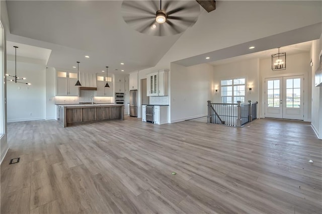 unfurnished living room featuring light wood-type flooring, sink, ceiling fan with notable chandelier, beverage cooler, and high vaulted ceiling