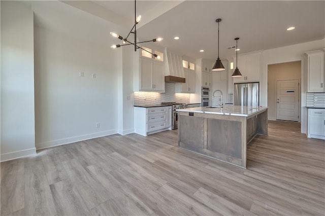 kitchen featuring appliances with stainless steel finishes, custom range hood, white cabinetry, pendant lighting, and a center island with sink