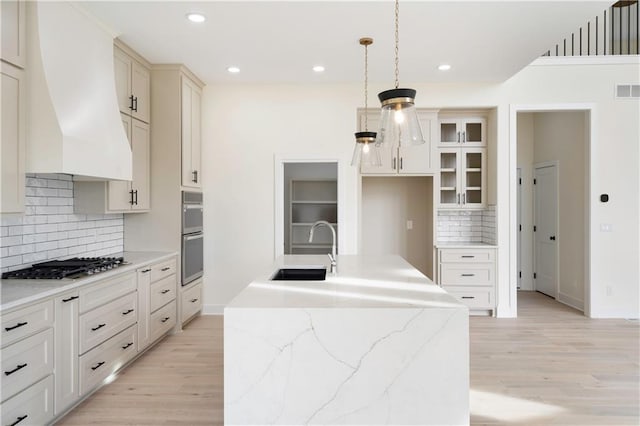 kitchen featuring sink, light wood-type flooring, pendant lighting, custom range hood, and a center island with sink