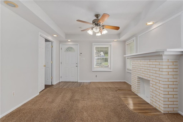 unfurnished living room featuring a raised ceiling, ceiling fan, a brick fireplace, and dark colored carpet