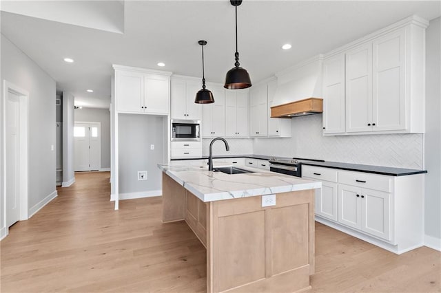 kitchen featuring premium range hood, pendant lighting, an island with sink, sink, and white cabinets