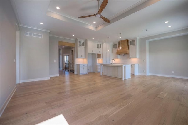 unfurnished living room featuring sink, ceiling fan, a tray ceiling, crown molding, and light wood-type flooring