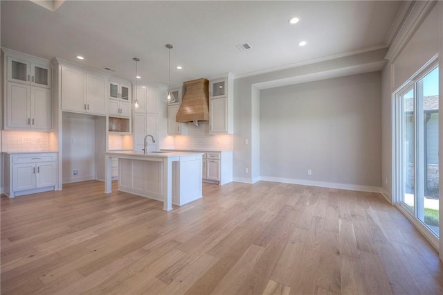 kitchen with white cabinetry, hanging light fixtures, a kitchen island with sink, crown molding, and custom range hood