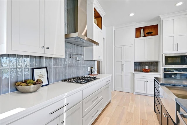 kitchen featuring wall chimney range hood, backsplash, appliances with stainless steel finishes, white cabinets, and light wood-type flooring