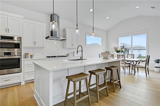 kitchen featuring an island with sink, hanging light fixtures, sink, white cabinets, and wall chimney range hood