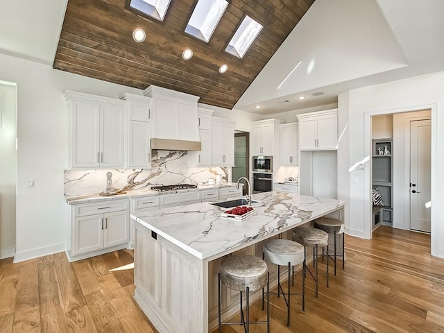 kitchen featuring a skylight, white cabinetry, light stone counters, backsplash, and a center island with sink