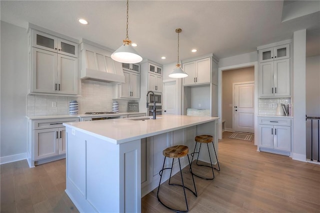 kitchen with custom exhaust hood, a kitchen island with sink, a sink, light countertops, and light wood-type flooring