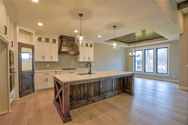 kitchen featuring a center island with sink, white cabinets, sink, custom exhaust hood, and light wood-type flooring