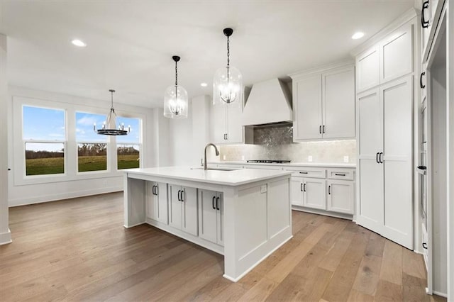 kitchen featuring premium range hood, a kitchen island with sink, white cabinetry, backsplash, and light hardwood / wood-style floors