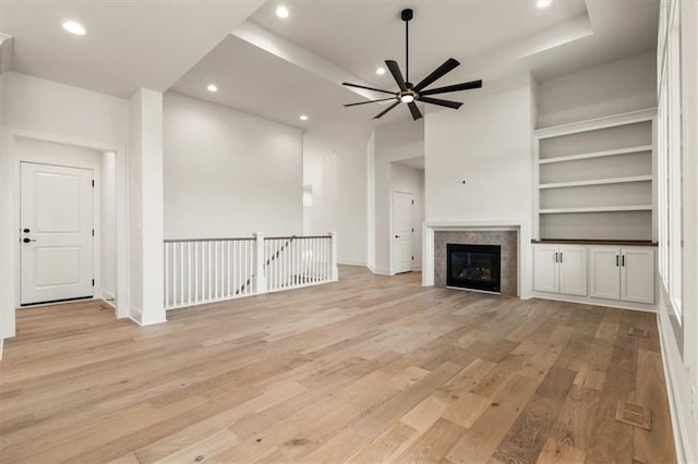 unfurnished living room with built in features, a raised ceiling, ceiling fan, and light wood-type flooring