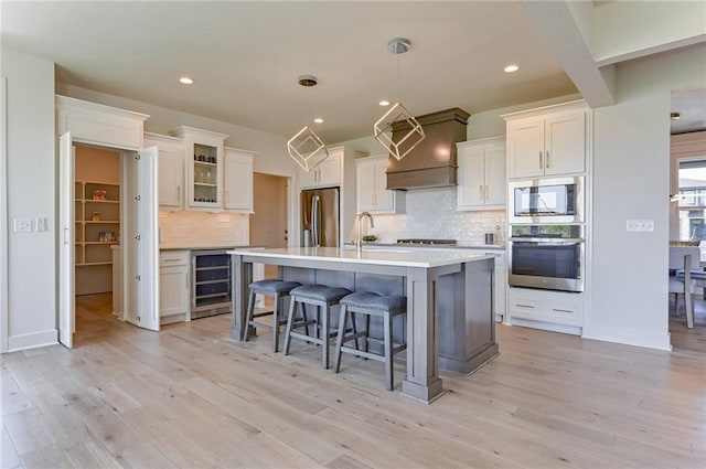 kitchen with appliances with stainless steel finishes, a kitchen island with sink, hanging light fixtures, white cabinets, and beverage cooler