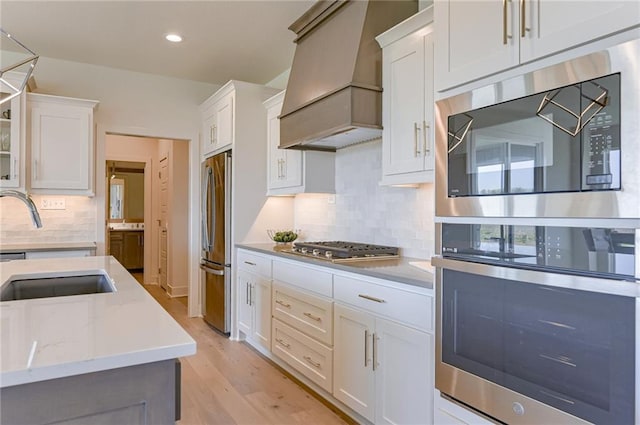 kitchen with white cabinetry, light wood-type flooring, premium range hood, appliances with stainless steel finishes, and decorative backsplash