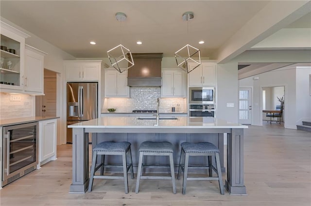 kitchen featuring appliances with stainless steel finishes, hanging light fixtures, premium range hood, light hardwood / wood-style floors, and beverage cooler