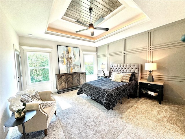 carpeted bedroom with a tray ceiling, multiple windows, a ceiling fan, and a decorative wall