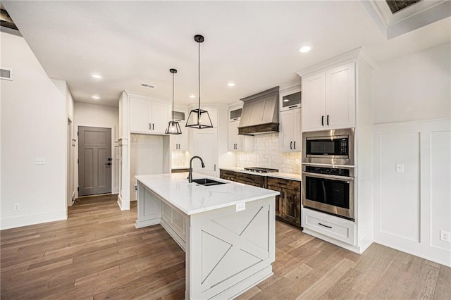 kitchen with a sink, appliances with stainless steel finishes, light wood-type flooring, custom exhaust hood, and backsplash