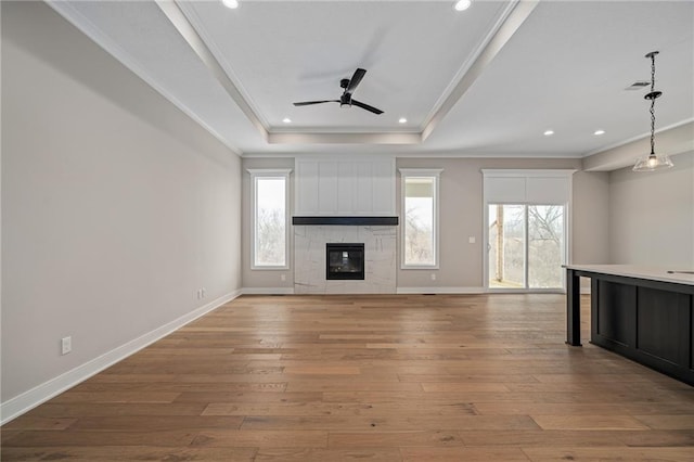 unfurnished living room featuring a tray ceiling, a large fireplace, light hardwood / wood-style floors, ceiling fan, and ornamental molding