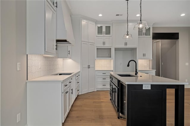 kitchen with a center island with sink, premium range hood, white cabinets, sink, and light wood-type flooring