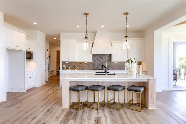 kitchen with light wood-type flooring, a center island with sink, custom range hood, and white cabinetry