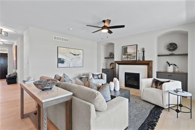 living room with ceiling fan, a tile fireplace, light wood-type flooring, and built in features