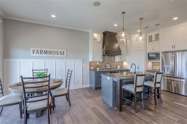 kitchen featuring pendant lighting, custom exhaust hood, white cabinets, an island with sink, and stainless steel appliances