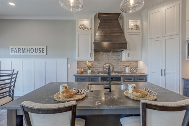 kitchen featuring custom exhaust hood, white cabinets, an island with sink, tasteful backsplash, and a breakfast bar area