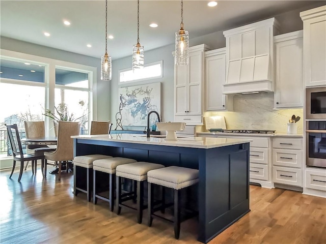 kitchen with white cabinetry, custom exhaust hood, decorative backsplash, light hardwood / wood-style flooring, and a center island with sink