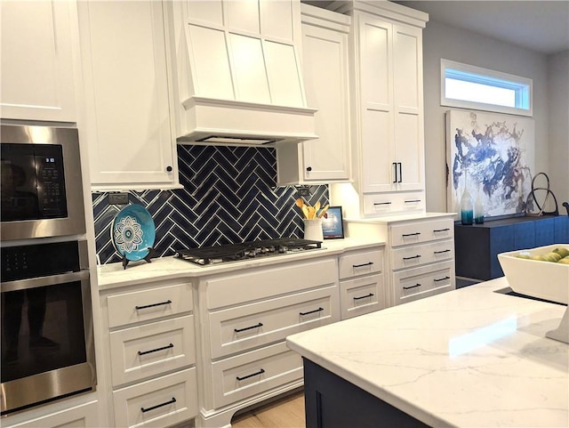 kitchen featuring backsplash, white cabinets, light stone counters, custom range hood, and stainless steel appliances