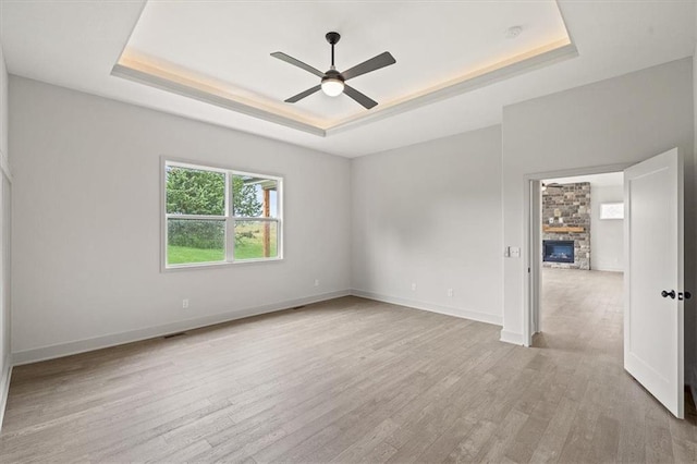 unfurnished room featuring ceiling fan, a stone fireplace, light hardwood / wood-style flooring, and a tray ceiling