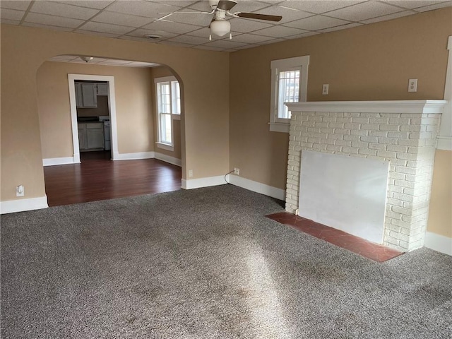 unfurnished room featuring a brick fireplace, a drop ceiling, dark carpet, and ceiling fan