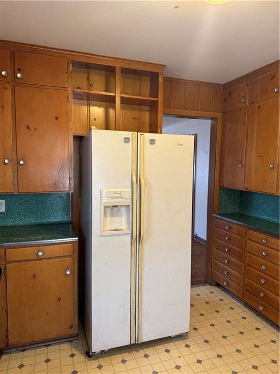 kitchen featuring dark countertops, light floors, brown cabinets, white refrigerator with ice dispenser, and open shelves