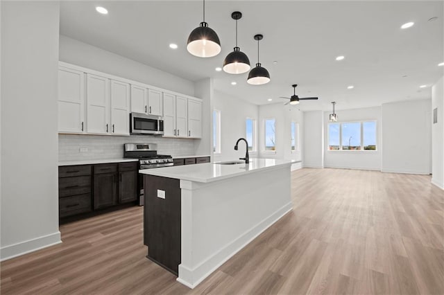 kitchen with a kitchen island with sink, stainless steel appliances, hanging light fixtures, white cabinets, and light hardwood / wood-style flooring