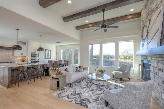 living room with ceiling fan, beamed ceiling, a stone fireplace, and light wood-type flooring