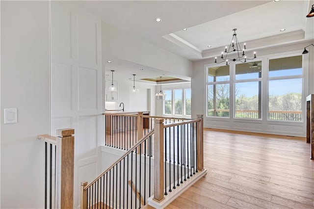 corridor with a tray ceiling, an upstairs landing, a notable chandelier, and light wood finished floors