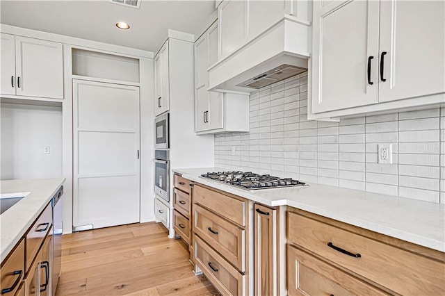 kitchen featuring custom exhaust hood, white cabinets, appliances with stainless steel finishes, light wood finished floors, and backsplash
