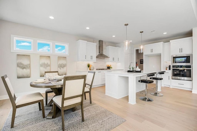 kitchen featuring wall chimney range hood, a kitchen island with sink, white cabinets, decorative light fixtures, and oven