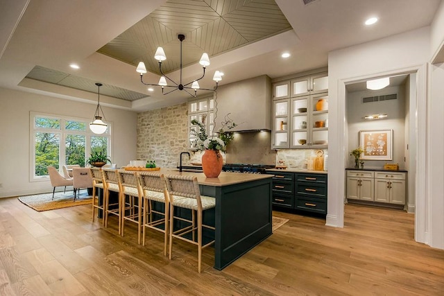 kitchen with light wood-type flooring, a kitchen breakfast bar, an island with sink, hanging light fixtures, and a tray ceiling