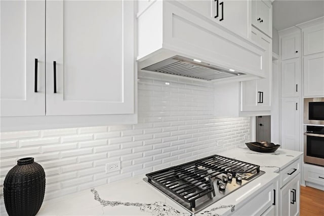 kitchen featuring appliances with stainless steel finishes, light stone counters, white cabinetry, backsplash, and premium range hood