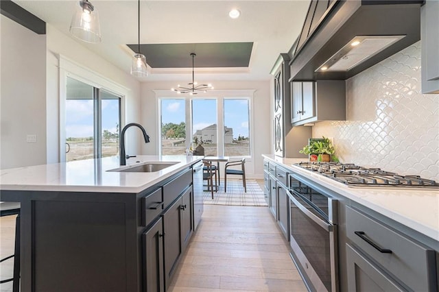 kitchen with a center island with sink, sink, hanging light fixtures, a tray ceiling, and stainless steel appliances