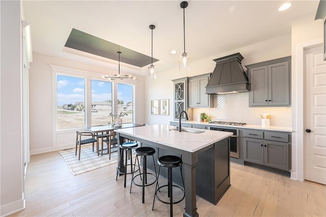 kitchen with premium range hood, stainless steel appliances, a tray ceiling, a kitchen island with sink, and sink