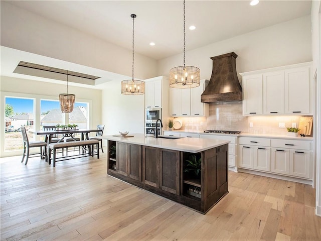 kitchen with pendant lighting, sink, white cabinets, a kitchen island with sink, and custom range hood