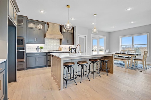 kitchen with appliances with stainless steel finishes, custom exhaust hood, a kitchen island with sink, sink, and hanging light fixtures