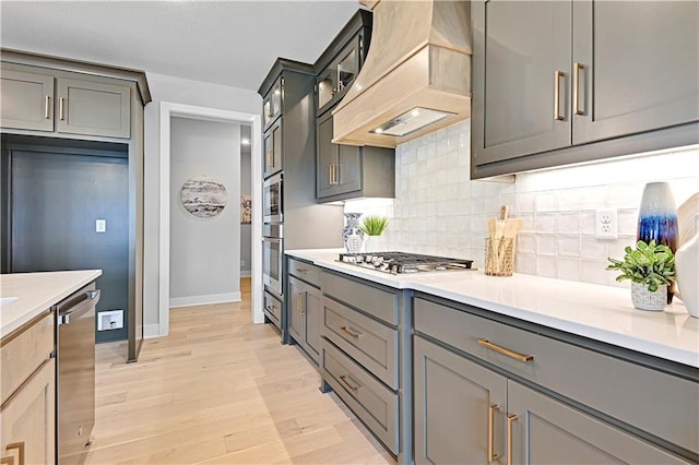 kitchen with custom exhaust hood, decorative backsplash, gray cabinets, light wood-type flooring, and appliances with stainless steel finishes