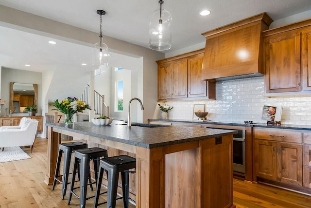 kitchen featuring premium range hood, sink, light wood-type flooring, a kitchen island with sink, and decorative backsplash