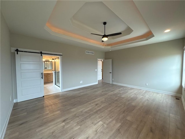 unfurnished bedroom featuring wood-type flooring, ceiling fan, a raised ceiling, a barn door, and ensuite bath