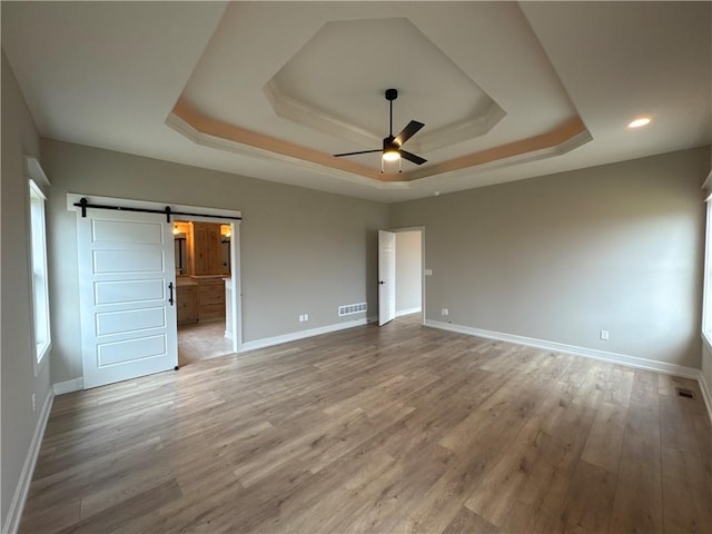 unfurnished bedroom with ceiling fan, ensuite bathroom, wood-type flooring, a raised ceiling, and a barn door