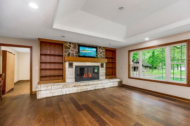 unfurnished living room featuring a stone fireplace, a raised ceiling, built in features, and dark wood-type flooring