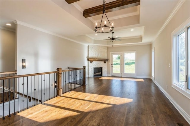 unfurnished living room featuring dark wood-type flooring, a tray ceiling, crown molding, a fireplace, and ceiling fan with notable chandelier